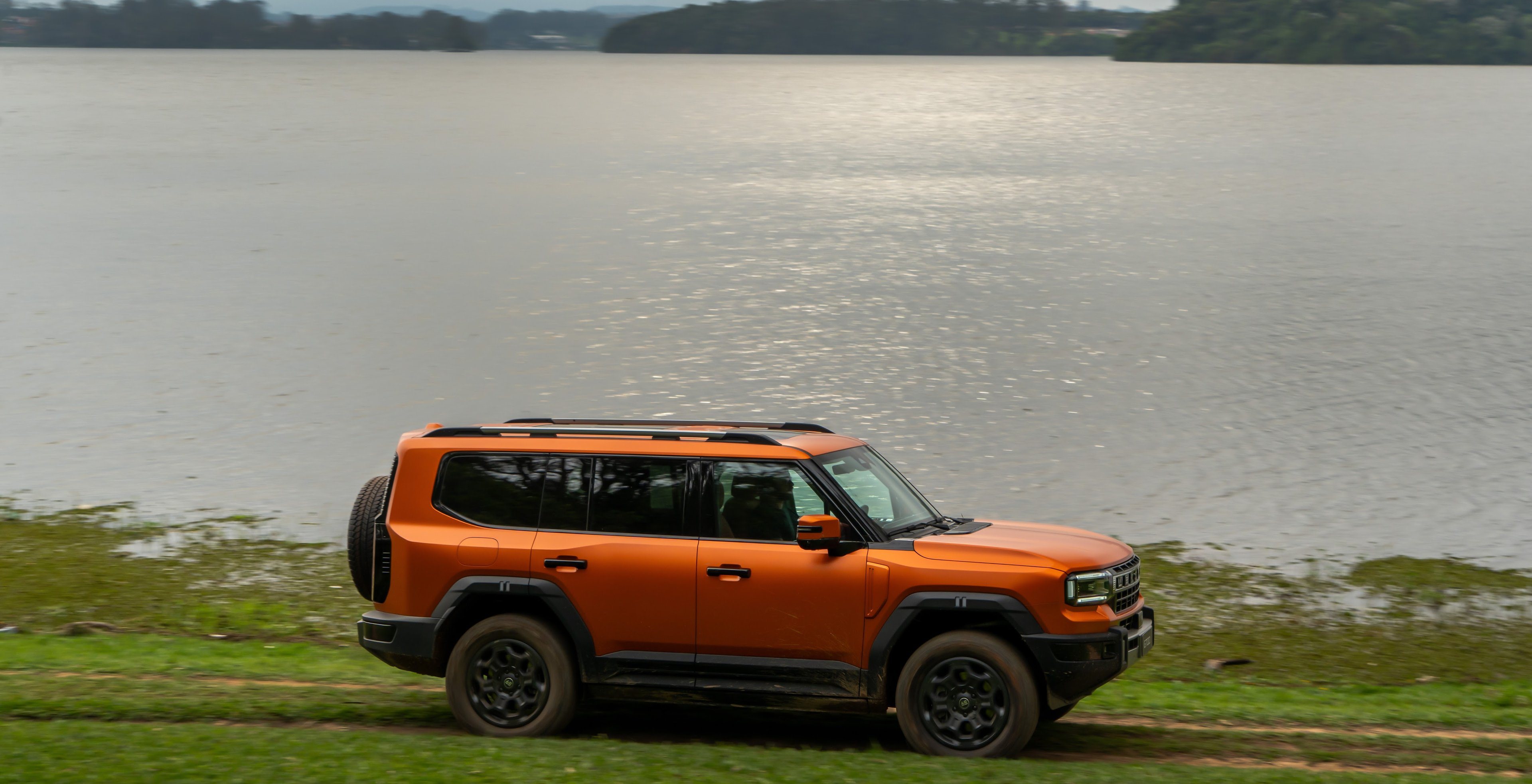 JETOUR G700 laranja de lado num gramado à beira de um lago, com céu nublado
