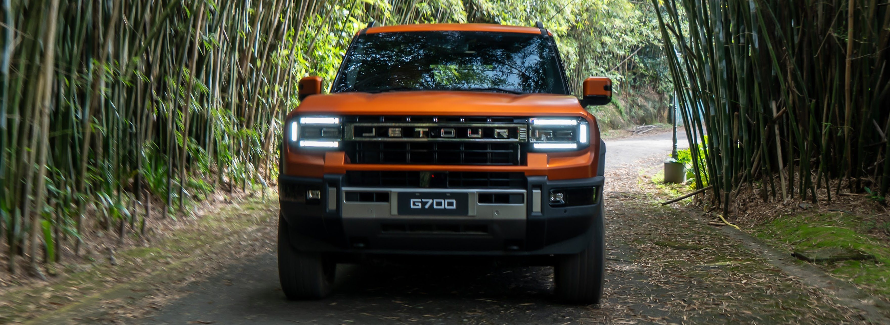 JETOUR G700 laranja de frente, numa estrada de terra dentro de um túnel de bambus