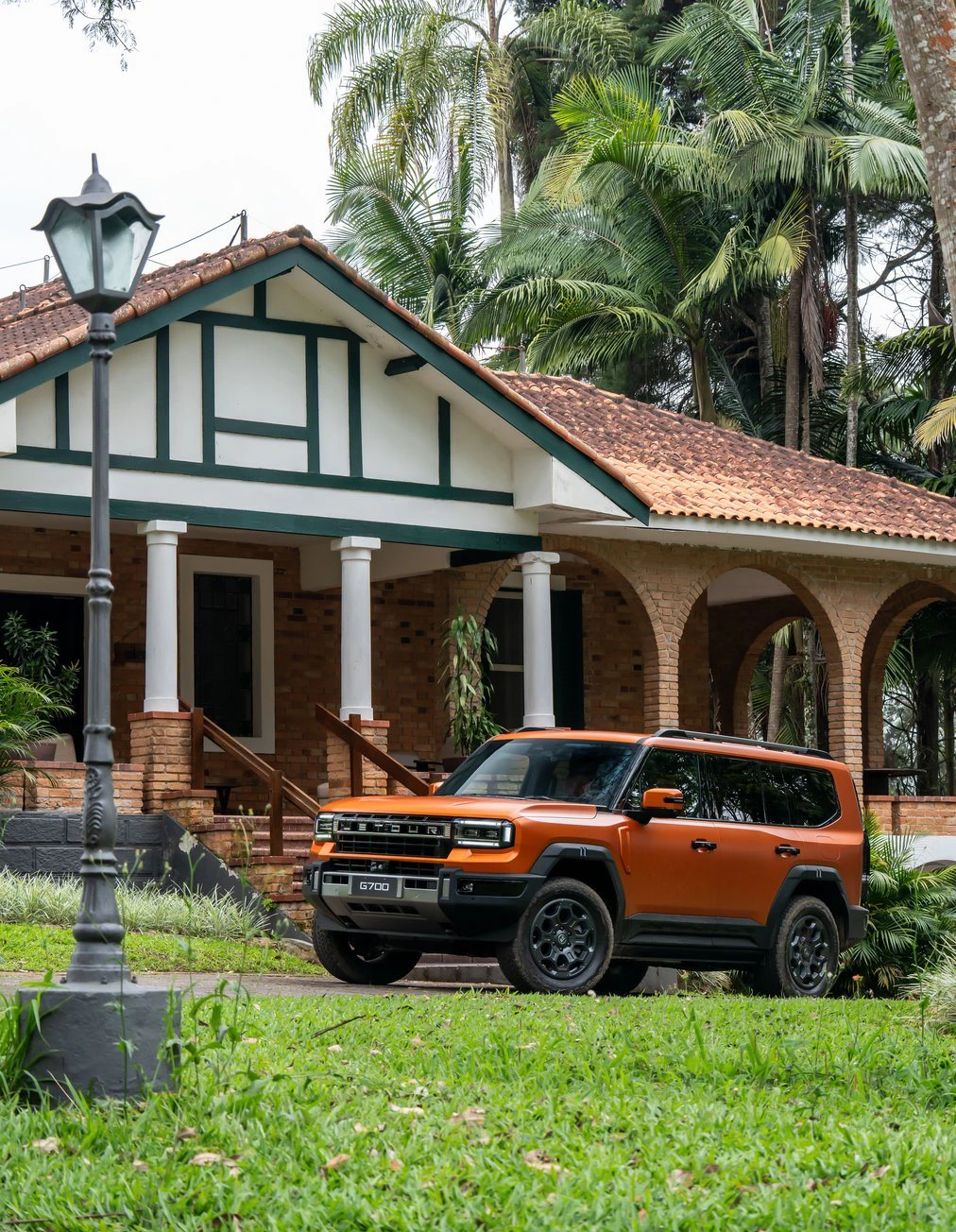 JETOUR G700 laranja diante de uma casa de tijolos com varanda, cercada de palmeiras