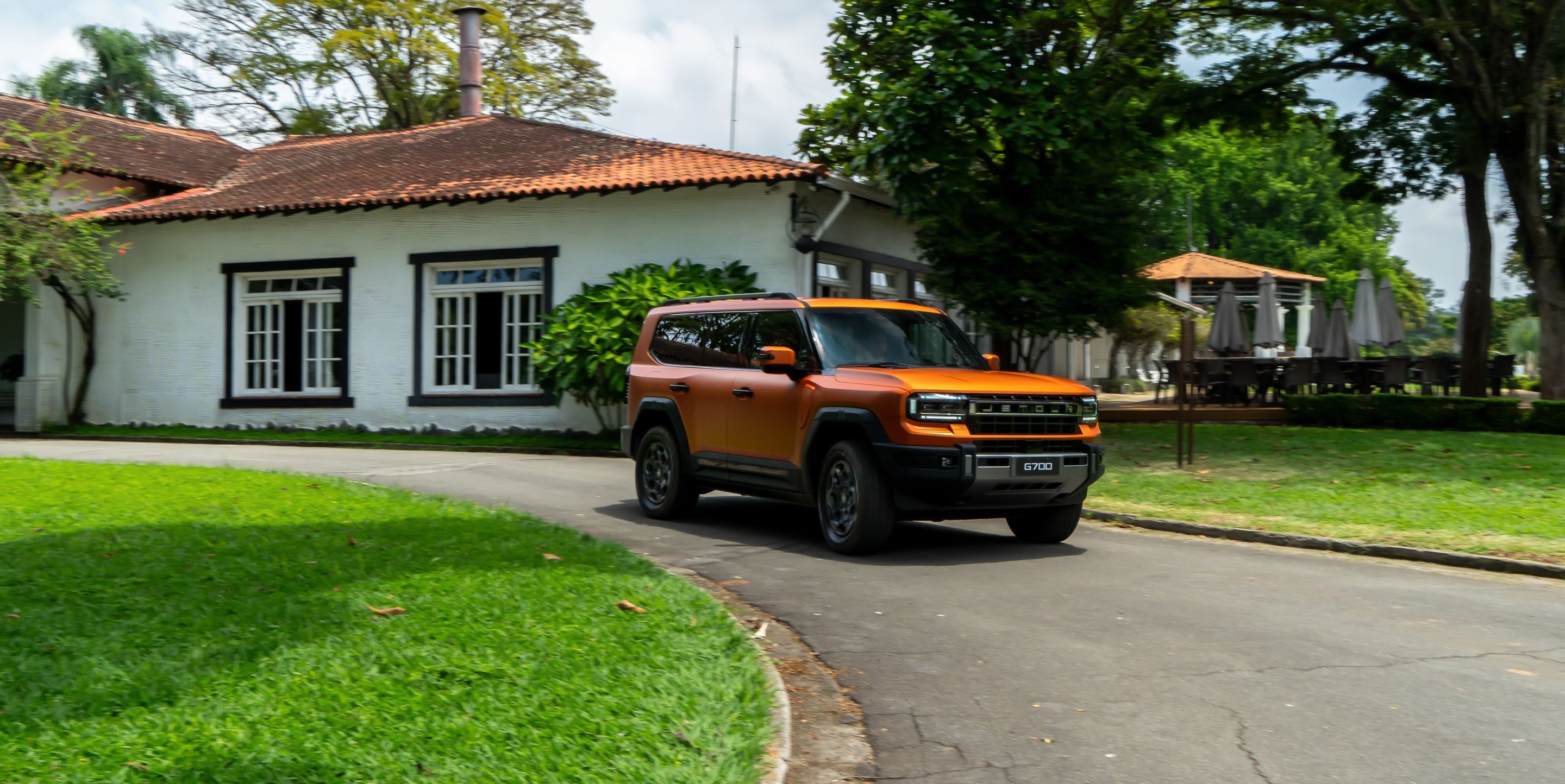 JETOUR G700 laranja numa estrada diante de uma casa branca de fazenda, com jardim ao redor